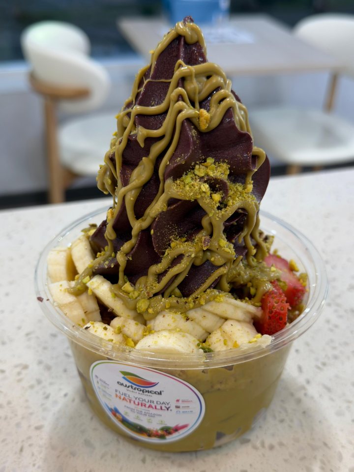A tall swirl of açaí soft serve with pistachio drizzle and crushed pistachios over banana and strawberries in a clear cup, on the counter at the Austropical café in Sydney, white chairs blurred behind.